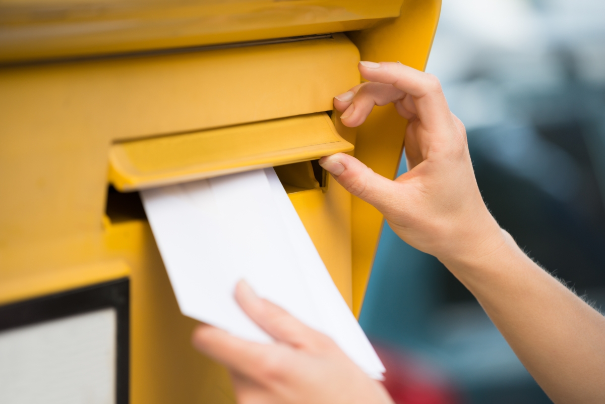 weibliche Hand steckt einen Briefumschlag in einen gelben Postbriefkasten